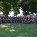 A large group of military personnel in camouflage uniforms pose for a formal unit photograph outdoors beneath large trees. The group consists of approximately 50-60 soldiers arranged in two to three rows, many holding rifles or wearing tactical gear. The setting appears to be a military installation or training area with tents and structures visible in the background.