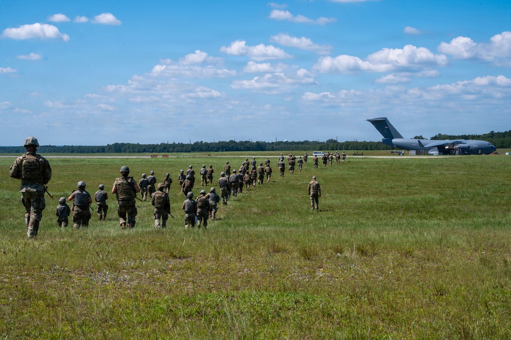 A column of military personnel in combat gear and helmets marches across an open grassy field toward a large military transport aircraft, likely a C-17 Globemaster III, with its rear cargo ramp open. The formation appears to be boarding or approaching the aircraft during what appears to be a training exercise or deployment operation. The scene takes place under a partly cloudy blue sky with a tree line visible in the background.