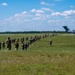 A column of military personnel in combat gear and helmets marches across an open grassy field toward a large military transport aircraft, likely a C-17 Globemaster III, with its rear cargo ramp open. The formation appears to be boarding or approaching the aircraft during what appears to be a training exercise or deployment operation. The scene takes place under a partly cloudy blue sky with a tree line visible in the background.