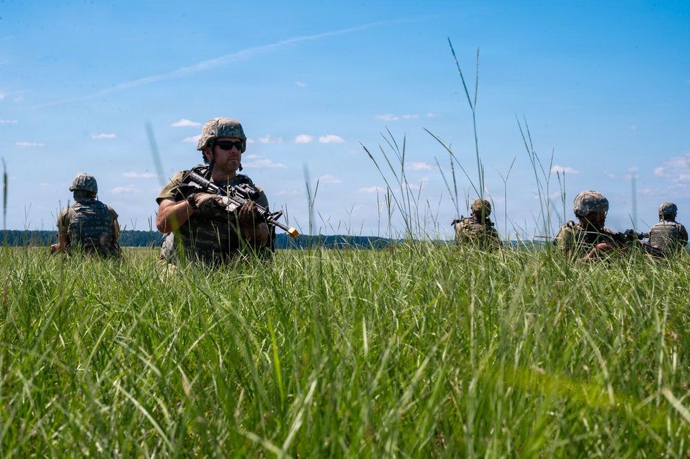 A group of military personnel in combat gear are positioned in a tall grass field, appearing to conduct a patrol or tactical exercise. The soldiers are equipped with rifles, body armor, and helmets, spread across the field in a dispersed formation. The setting appears to be a training environment during daytime under clear skies.