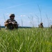 A group of military personnel in combat gear are positioned in a tall grass field, appearing to conduct a patrol or tactical exercise. The soldiers are equipped with rifles, body armor, and helmets, spread across the field in a dispersed formation. The setting appears to be a training environment during daytime under clear skies.