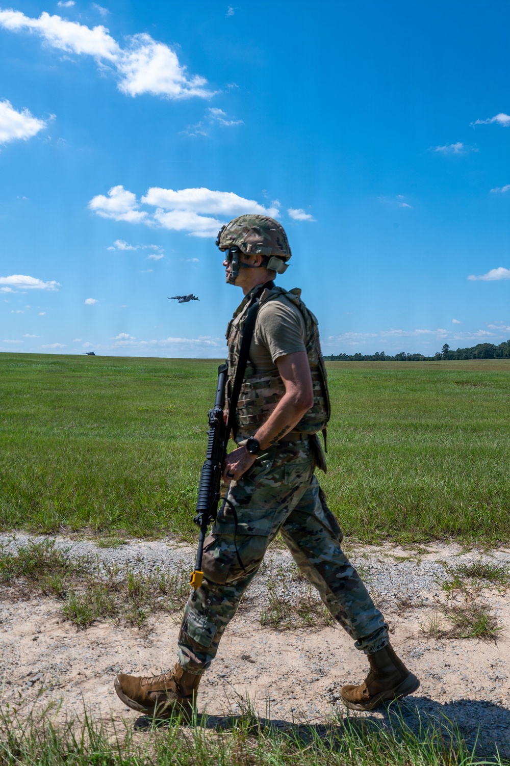 A uniformed military service member in full combat gear walks along a gravel path on what appears to be a training field or airfield. In the background, a military aircraft (likely a C-130 or similar transport) is visible in flight over an open grassy field. The image appears to document a military training exercise under clear daytime conditions.