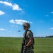 A uniformed military service member in full combat gear walks along a gravel path on what appears to be a training field or airfield. In the background, a military aircraft (likely a C-130 or similar transport) is visible in flight over an open grassy field. The image appears to document a military training exercise under clear daytime conditions.