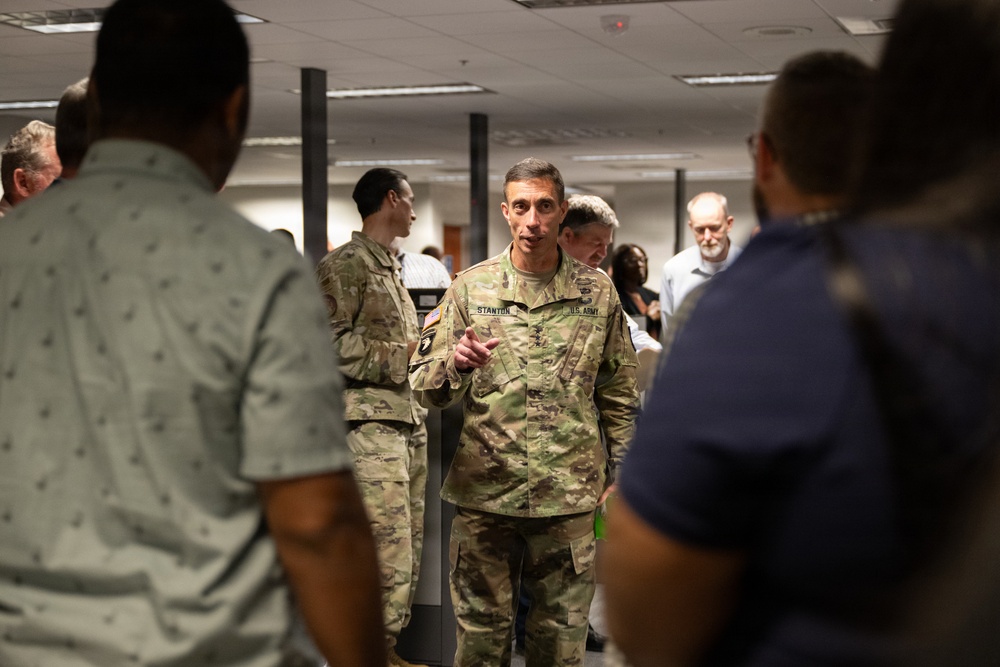 A U.S. Army soldier with the name tape 'STANTON' and 'U.S. ARMY' visible on their OCP (Operational Camouflage Pattern) uniform is speaking and gesturing to a group of people in what appears to be an indoor office or briefing room. The soldier appears to be addressing a mixed group of military and civilian personnel. The setting suggests an official briefing, tour, or meeting event.