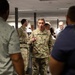 A U.S. Army soldier with the name tape 'STANTON' and 'U.S. ARMY' visible on their OCP (Operational Camouflage Pattern) uniform is speaking and gesturing to a group of people in what appears to be an indoor office or briefing room. The soldier appears to be addressing a mixed group of military and civilian personnel. The setting suggests an official briefing, tour, or meeting event.