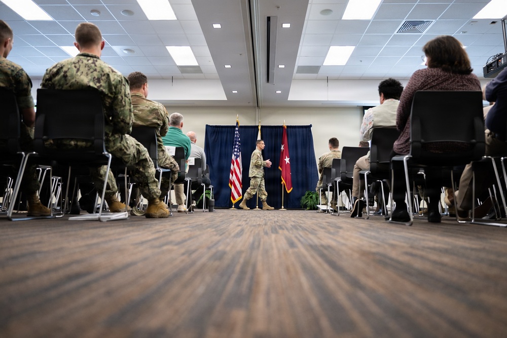 A military briefing or ceremony is taking place in a conference room, with an audience of uniformed military personnel and civilians seated in rows of chairs. A single soldier in Army combat uniform is walking near a podium area flanked by an American flag and what appears to be a general officer's flag (red with white stars). The low-angle perspective from floor level creates a dramatic compositional view of the event.