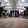 Lt. Gen. Stanton at DISA HQ. A military briefing or ceremony is taking place in a conference room, with an audience of uniformed military personnel and civilians seated in rows of chairs. A single soldier in Army combat uniform is walking near a podium area flanked by an American flag and what appears to be a general officer's flag (red with white stars). The low-angle perspective from floor level creates a dramatic compositional view of the event.