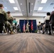A military briefing or ceremony is taking place in a conference room, with an audience of uniformed military personnel and civilians seated in rows of chairs. A single soldier in Army combat uniform is walking near a podium area flanked by an American flag and what appears to be a general officer's flag (red with white stars). The low-angle perspective from floor level creates a dramatic compositional view of the event.