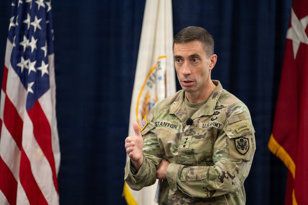 A U.S. Army officer wearing the rank of Lieutenant General (three stars) and identified by name tape as 'Stanton' is speaking at an official military event. He is dressed in Army Combat Uniform (ACU) with MultiCam pattern and displays a Ranger tab and U.S. Army shoulder patch. Multiple flags, including the U.S. flag and a military organizational flag, are visible in the background.