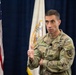 A U.S. Army officer wearing the rank of Lieutenant General (three stars) and identified by name tape as 'Stanton' is speaking at an official military event. He is dressed in Army Combat Uniform (ACU) with MultiCam pattern and displays a Ranger tab and U.S. Army shoulder patch. Multiple flags, including the U.S. flag and a military organizational flag, are visible in the background.