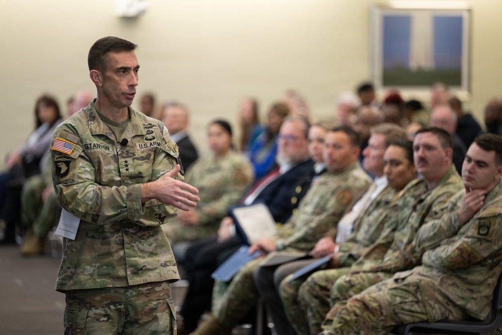 A U.S. Army officer with the name tape 'STANTON' and three stars on his collar is addressing a mixed audience of military personnel and civilians in what appears to be a briefing or town hall setting. The speaker wears an OCP (Operational Camouflage Pattern) uniform with a 101st Airborne Division patch and a U.S. flag patch on the left shoulder. The audience is seated in rows and appears attentive.