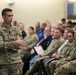 A U.S. Army officer with the name tape 'STANTON' and three stars on his collar is addressing a mixed audience of military personnel and civilians in what appears to be a briefing or town hall setting. The speaker wears an OCP (Operational Camouflage Pattern) uniform with a 101st Airborne Division patch and a U.S. flag patch on the left shoulder. The audience is seated in rows and appears attentive.
