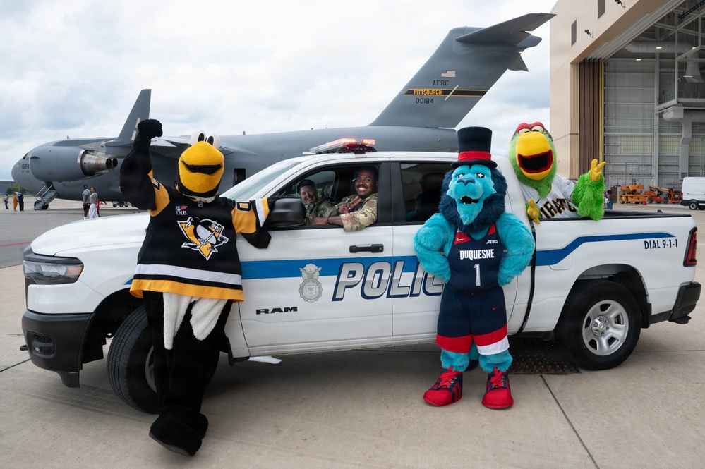 Three sports mascots pose alongside a white RAM police pickup truck on a military airfield tarmac, with a large military cargo aircraft visible in the background. Two individuals in military uniforms are seated inside the police vehicle. The scene appears to be a community outreach or promotional event combining local Pittsburgh sports teams with law enforcement and military presence.