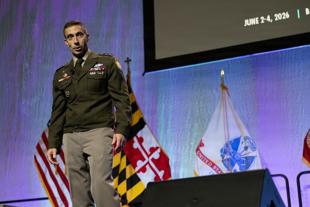 A U.S. Army officer in dress green uniform is speaking on a stage at what appears to be a military conference or symposium. Behind him are multiple flags including the U.S. flag, the Maryland state flag, and a United States Army flag. A projection screen in the background displays the dates 'June 2-4, 2026'.