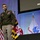 Lt. Gen. Stanton speaks at AFCEA TNC 26 A U.S. Army officer in dress green uniform is speaking on a stage at what appears to be a military conference or symposium. Behind him are multiple flags including the U.S. flag, the Maryland state flag, and a United States Army flag. A projection screen in the background displays the dates 'June 2-4, 2026'.