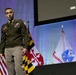 A U.S. Army officer in dress green uniform is speaking on a stage at what appears to be a military conference or symposium. Behind him are multiple flags including the U.S. flag, the Maryland state flag, and a United States Army flag. A projection screen in the background displays the dates 'June 2-4, 2026'.
