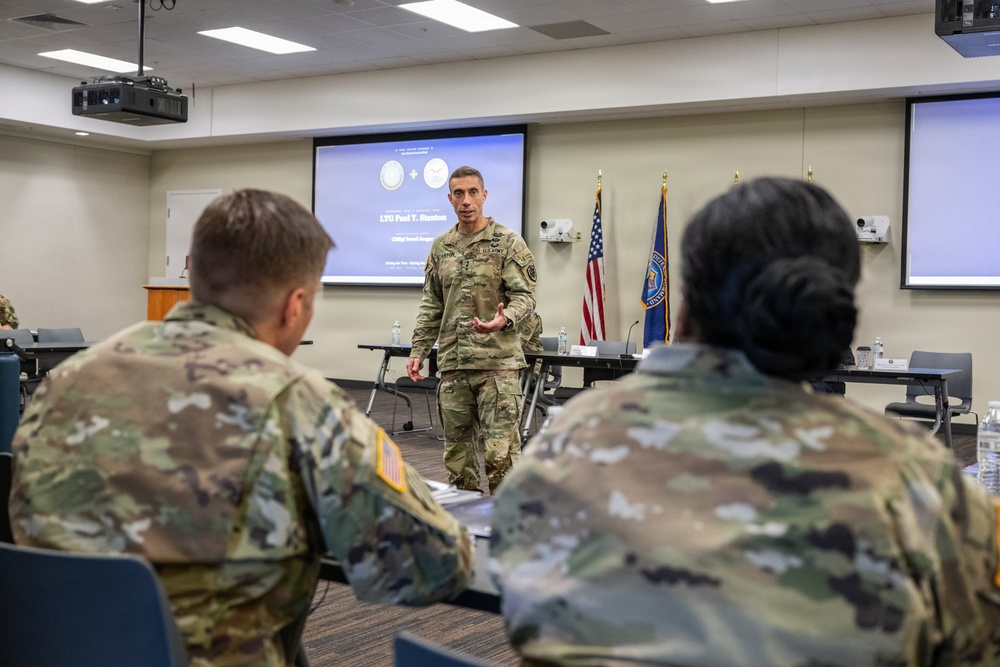 A U.S. Army officer in OCP (Operational Camouflage Pattern) uniform is presenting to a group of seated military personnel in what appears to be a briefing or classroom setting. A projection screen behind the speaker displays a slide referencing 'LTG Paul T. Stanton' and what appear to be two military insignia seals. The setting suggests a formal military briefing or training event.