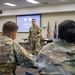 A U.S. Army officer in OCP (Operational Camouflage Pattern) uniform is presenting to a group of seated military personnel in what appears to be a briefing or classroom setting. A projection screen behind the speaker displays a slide referencing 'LTG Paul T. Stanton' and what appear to be two military insignia seals. The setting suggests a formal military briefing or training event.