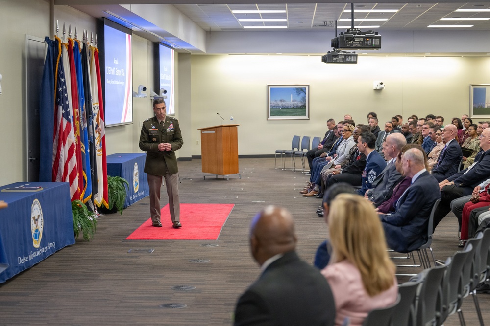 A military officer in Army dress uniform is delivering a speech or presentation at a formal event, standing on a red carpet in front of a seated audience. The setting appears to be an official government or defense agency conference room, identifiable by the 'Defense Information Systems Agency' (DISA) branded table covering and multiple military flags. A projection screen displays a slide behind a wooden podium.