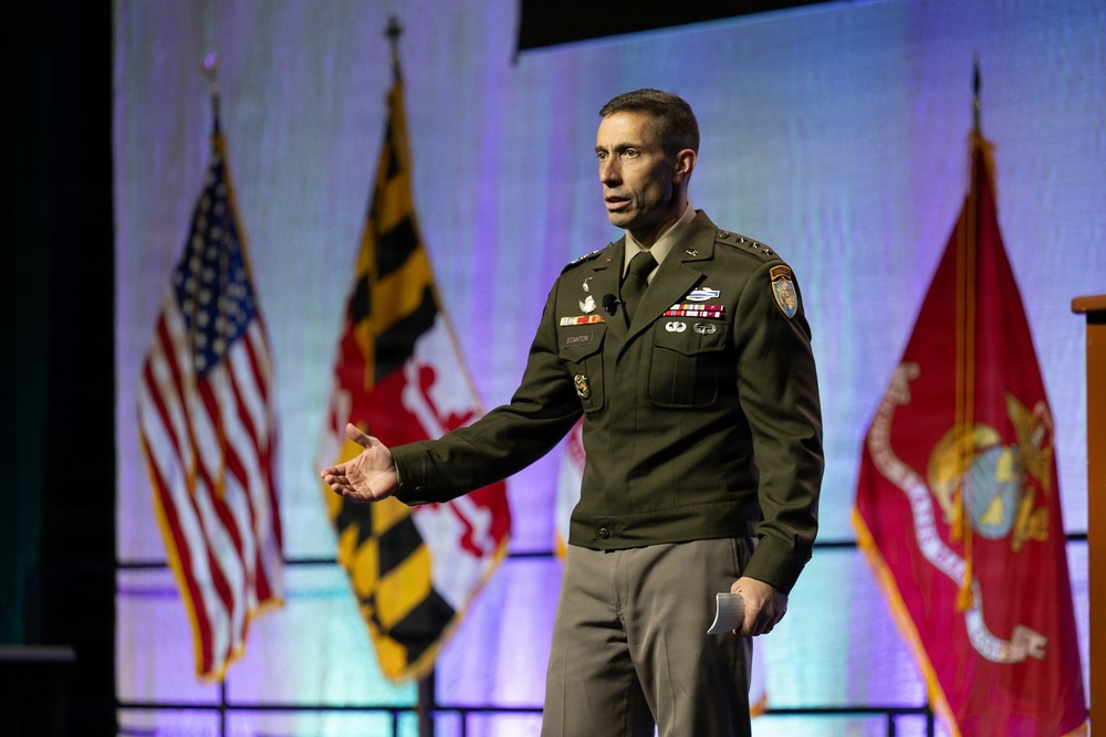 A U.S. Army officer in dress green uniform is delivering a speech or presentation on a stage. He is gesturing with his right hand while holding papers in his left hand. Multiple flags are visible in the background, including the U.S. flag, Maryland state flag, and U.S. Marine Corps flag.
