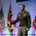 Lt. Gen. Stanton speaks at AFCEA TNC A U.S. Army officer in dress green uniform is delivering a speech or presentation on a stage. He is gesturing with his right hand while holding papers in his left hand. Multiple flags are visible in the background, including the U.S. flag, Maryland state flag, and U.S. Marine Corps flag.