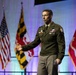 A U.S. Army officer in dress green uniform is delivering a speech or presentation on a stage. He is gesturing with his right hand while holding papers in his left hand. Multiple flags are visible in the background, including the U.S. flag, Maryland state flag, and U.S. Marine Corps flag.