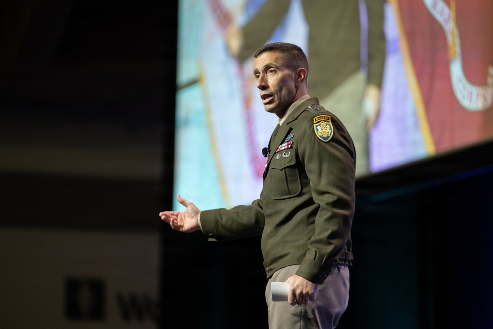 A U.S. Army officer in dress green uniform is delivering a presentation or speech on a stage. He is gesturing with his right hand while holding papers in his left, with a large projection screen visible behind him. The setting appears to be a formal conference or military briefing event.