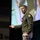 Lt. Gen. Stanton speaks at AFCEA Tech Net Cyber A U.S. Army officer in dress green uniform is delivering a presentation or speech on a stage. He is gesturing with his right hand while holding papers in his left, with a large projection screen visible behind him. The setting appears to be a formal conference or military briefing event.