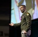 A U.S. Army officer in dress green uniform is delivering a presentation or speech on a stage. He is gesturing with his right hand while holding papers in his left, with a large projection screen visible behind him. The setting appears to be a formal conference or military briefing event.