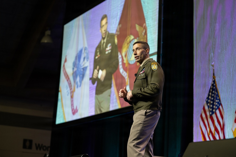 A military officer in green Army dress uniform is delivering a presentation on stage at what appears to be a professional conference or symposium. Behind him, a large projection screen displays an image of another military officer standing in front of military flags. A partial US flag is visible on the right side of the stage.