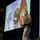 Lt. Gen. Stanton speaks at AFCEA Tech Net Cyber A military officer in green Army dress uniform is delivering a presentation on stage at what appears to be a professional conference or symposium. Behind him, a large projection screen displays an image of another military officer standing in front of military flags. A partial US flag is visible on the right side of the stage.