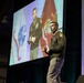 A military officer in green Army dress uniform is delivering a presentation on stage at what appears to be a professional conference or symposium. Behind him, a large projection screen displays an image of another military officer standing in front of military flags. A partial US flag is visible on the right side of the stage.