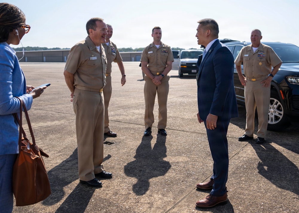 A formal outdoor meeting on a military installation tarmac or parking area, showing U.S. Navy personnel in tan service uniforms greeting a civilian official in a navy blue suit. The interaction appears cordial, with the primary naval officer and civilian engaged in conversation while other uniformed personnel stand at ease in the background. Government vehicles are visible in the background, suggesting an official arrival or reception event.