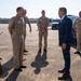 A formal outdoor meeting on a military installation tarmac or parking area, showing U.S. Navy personnel in tan service uniforms greeting a civilian official in a navy blue suit. The interaction appears cordial, with the primary naval officer and civilian engaged in conversation while other uniformed personnel stand at ease in the background. Government vehicles are visible in the background, suggesting an official arrival or reception event.