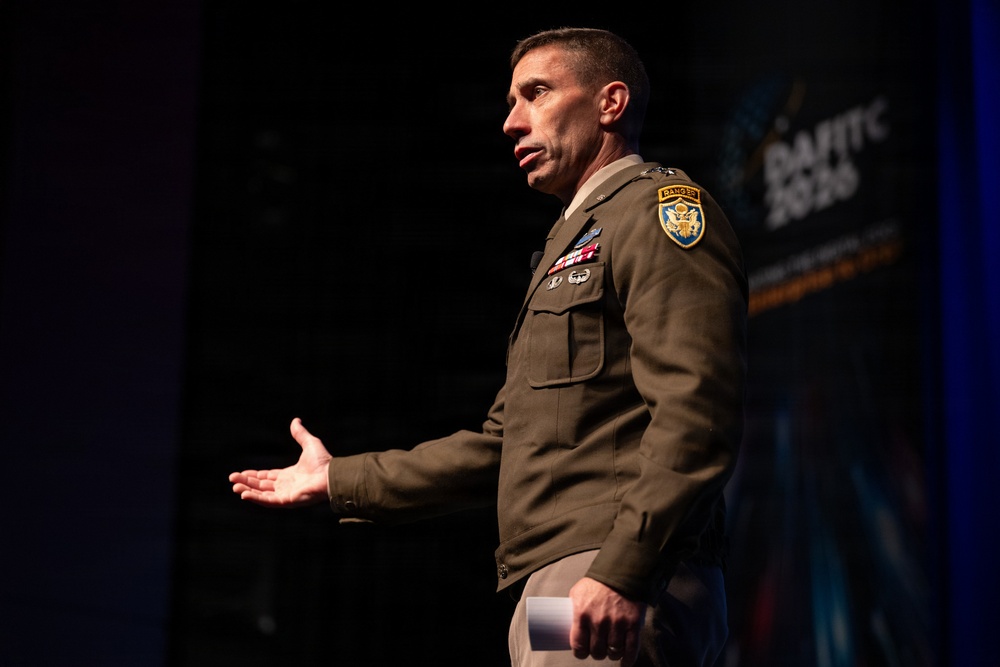 A U.S. Army officer in Army Green Service Uniform is speaking at what appears to be a military or defense technology conference, identifiable by the 'DAVITC 2026' banner visible in the background. The officer is gesturing with an open hand, suggesting active engagement with an audience. Multiple military insignia, including a Ranger tab and shoulder patch, are visible on the uniform.