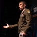 A U.S. Army officer in Army Green Service Uniform is speaking at what appears to be a military or defense technology conference, identifiable by the 'DAVITC 2026' banner visible in the background. The officer is gesturing with an open hand, suggesting active engagement with an audience. Multiple military insignia, including a Ranger tab and shoulder patch, are visible on the uniform.