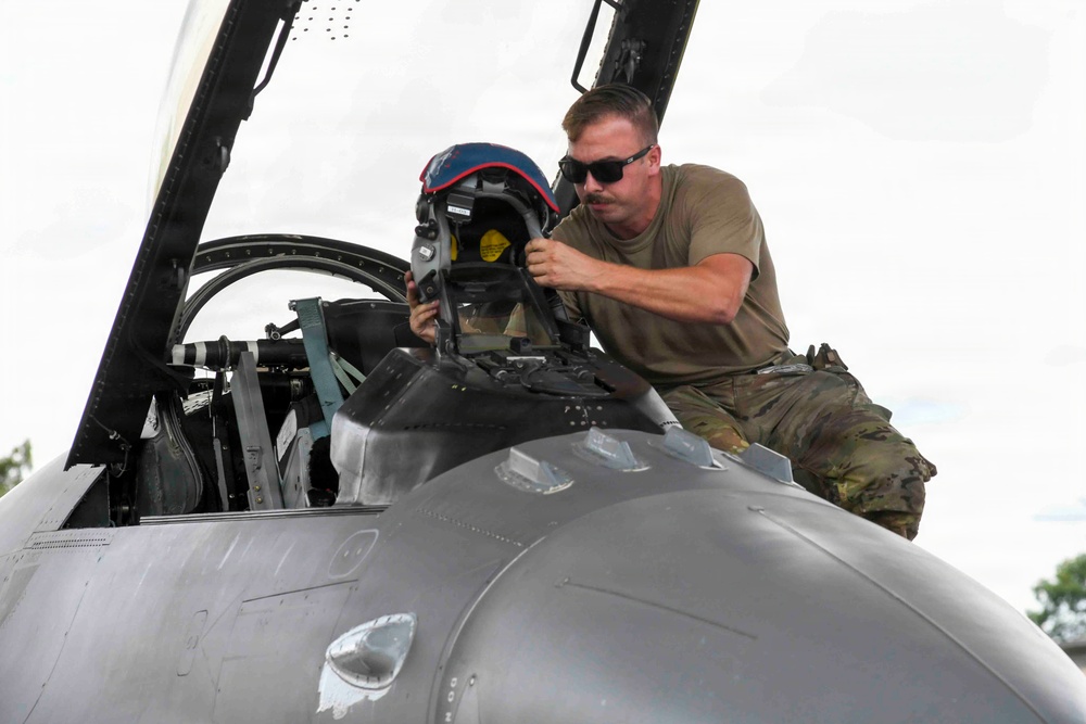 A military crew chief or ground technician in an olive drab t-shirt and multicam trousers is working on the cockpit of a military jet aircraft, handling a pilot's flight helmet. The aircraft appears to be a single-seat fighter jet, likely an F-16 Fighting Falcon, based on the cockpit configuration and fuselage design. The work appears to be pre-flight or post-flight equipment handling on a flight line.