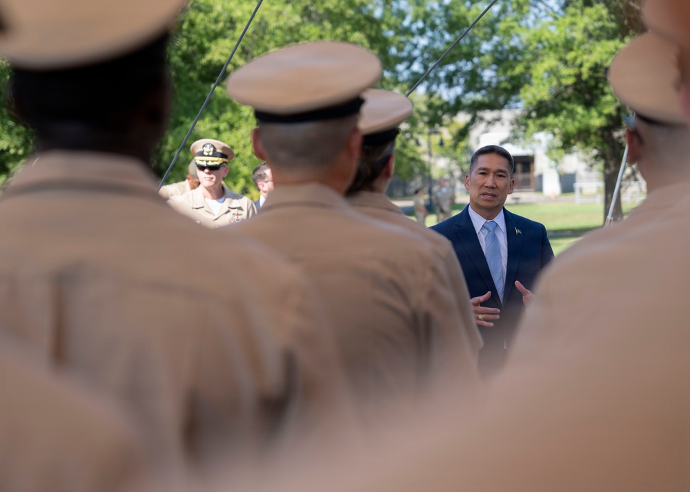 A civilian official in a dark suit and light blue tie appears to be addressing a formation of military personnel in tan/khaki uniforms from behind. A senior naval officer wearing sunglasses and a white uniform with gold insignia is visible in the background to the left. The setting appears to be an outdoor military ceremony or review on a sunny day.