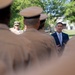 A civilian official in a dark suit and light blue tie appears to be addressing a formation of military personnel in tan/khaki uniforms from behind. A senior naval officer wearing sunglasses and a white uniform with gold insignia is visible in the background to the left. The setting appears to be an outdoor military ceremony or review on a sunny day.