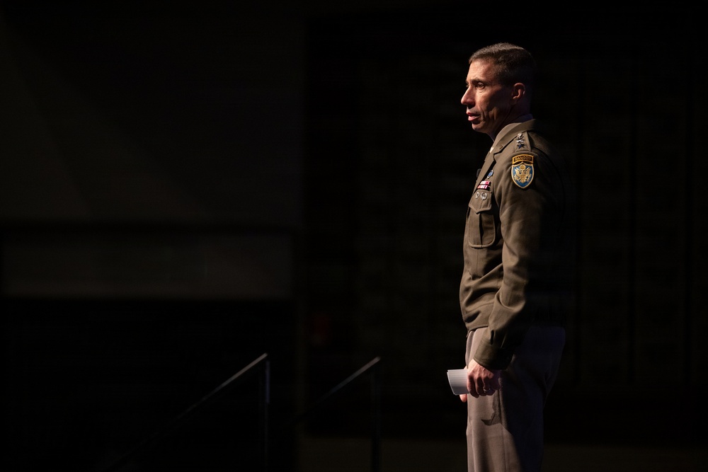 A male military officer in olive/khaki dress uniform is photographed in profile against a dark background, appearing to be speaking or presenting at a formal event. He is holding what appears to be a small piece of paper or note card. The dramatic side lighting creates a high-contrast, cinematic composition.