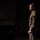 Lt. Gen. Stanton Speaks at DAFITC A male military officer in olive/khaki dress uniform is photographed in profile against a dark background, appearing to be speaking or presenting at a formal event. He is holding what appears to be a small piece of paper or note card. The dramatic side lighting creates a high-contrast, cinematic composition.