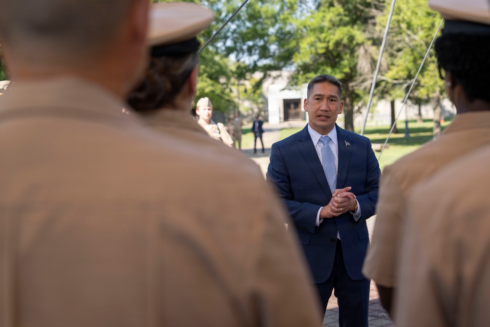 A man in a navy blue suit with a light blue tie stands facing a group of uniformed military personnel in tan/khaki uniforms. He appears to be addressing or speaking to the service members in an outdoor setting with trees and a building visible in the background. The composition suggests a formal military or official civilian-military engagement.
