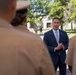 A man in a navy blue suit with a light blue tie stands facing a group of uniformed military personnel in tan/khaki uniforms. He appears to be addressing or speaking to the service members in an outdoor setting with trees and a building visible in the background. The composition suggests a formal military or official civilian-military engagement.