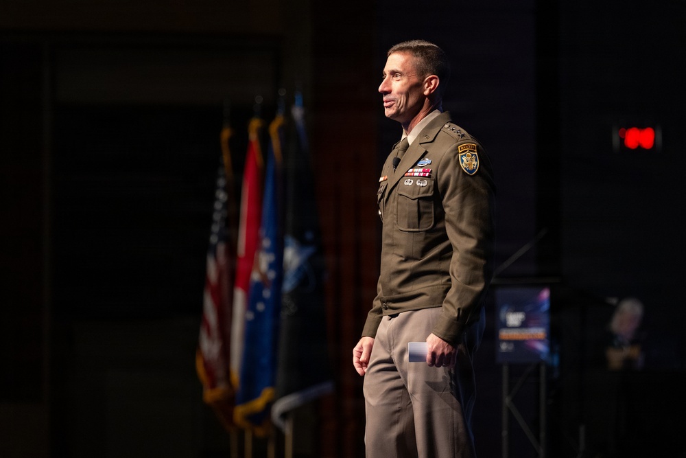 A senior U.S. Army officer in dress uniform stands on a stage, appearing to address an audience at a formal military or defense-related event. He is wearing an olive green Army Service Uniform with visible rank insignia, unit patches, and service ribbons. Multiple flags, including the U.S. flag, are displayed in the background.