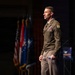 A senior U.S. Army officer in dress uniform stands on a stage, appearing to address an audience at a formal military or defense-related event. He is wearing an olive green Army Service Uniform with visible rank insignia, unit patches, and service ribbons. Multiple flags, including the U.S. flag, are displayed in the background.