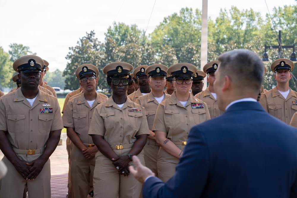 A group of U.S. Navy Chief Petty Officers in service khaki uniforms stand at attention while being addressed by a civilian in a dark blue suit. The formation appears to be an outdoor ceremony or briefing taking place on a military installation. The personnel display various service ribbons and rank insignia consistent with Chief Petty Officer grades.