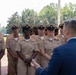 A group of U.S. Navy Chief Petty Officers in service khaki uniforms stand at attention while being addressed by a civilian in a dark blue suit. The formation appears to be an outdoor ceremony or briefing taking place on a military installation. The personnel display various service ribbons and rank insignia consistent with Chief Petty Officer grades.