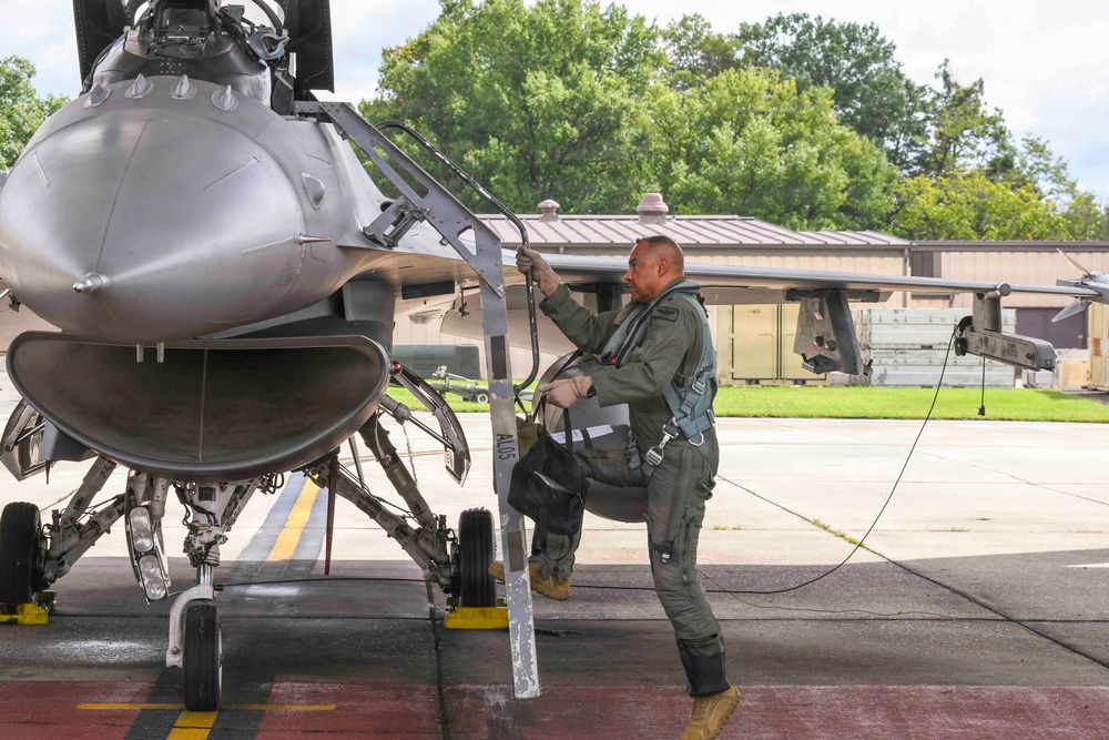 A military pilot in full flight gear is climbing a boarding ladder to access an F-16 Fighting Falcon jet aircraft on a flight line apron. The pilot is wearing an olive green flight suit with harness and carrying a flight bag. The setting appears to be a military air base with hangars and trees visible in the background.