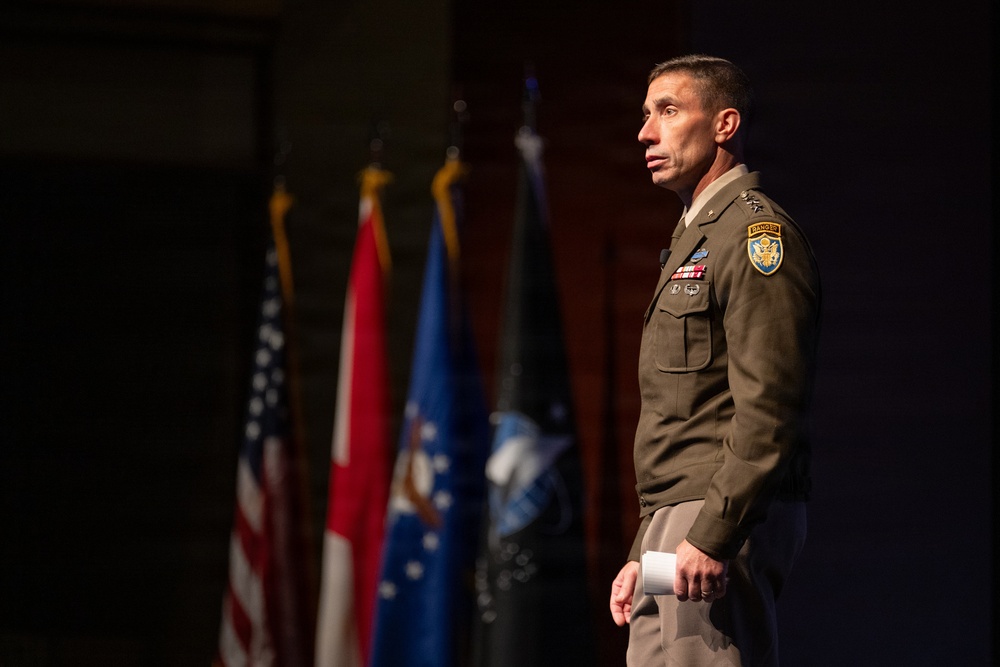 A U.S. Army officer in dress uniform stands on a stage, appearing to deliver a speech or presentation. He is wearing an olive green Army Service Uniform with visible rank insignia, a Ranger tab, a shoulder patch, and service ribbons. Multiple flags are displayed in the background, including the U.S. flag and what appear to be military branch and unit flags.