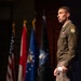 A U.S. Army officer in dress uniform stands on a stage, appearing to deliver a speech or presentation. He is wearing an olive green Army Service Uniform with visible rank insignia, a Ranger tab, a shoulder patch, and service ribbons. Multiple flags are displayed in the background, including the U.S. flag and what appear to be military branch and unit flags.